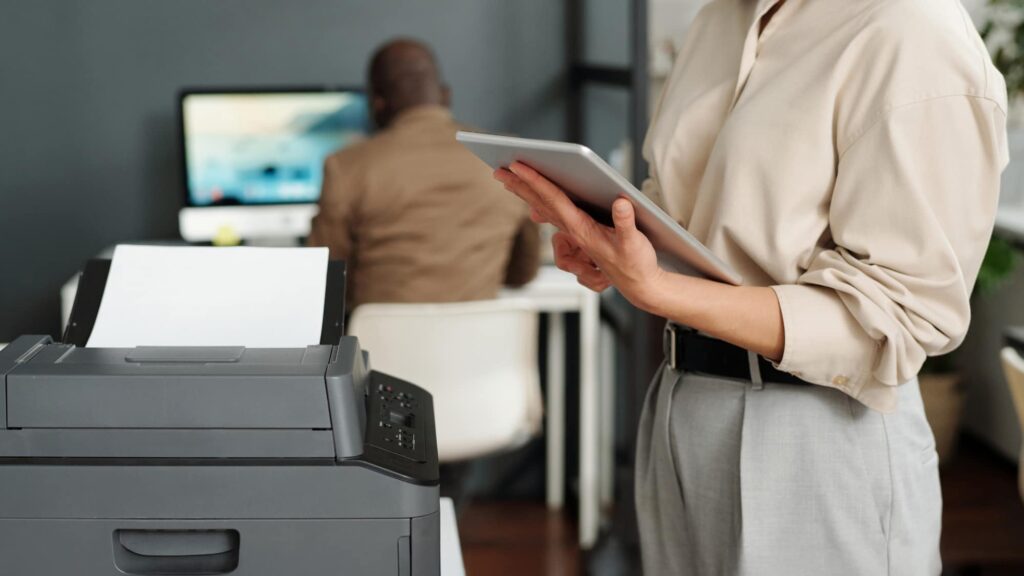 Employee using inventory management system on tablet next to office printer for tracking and service management.