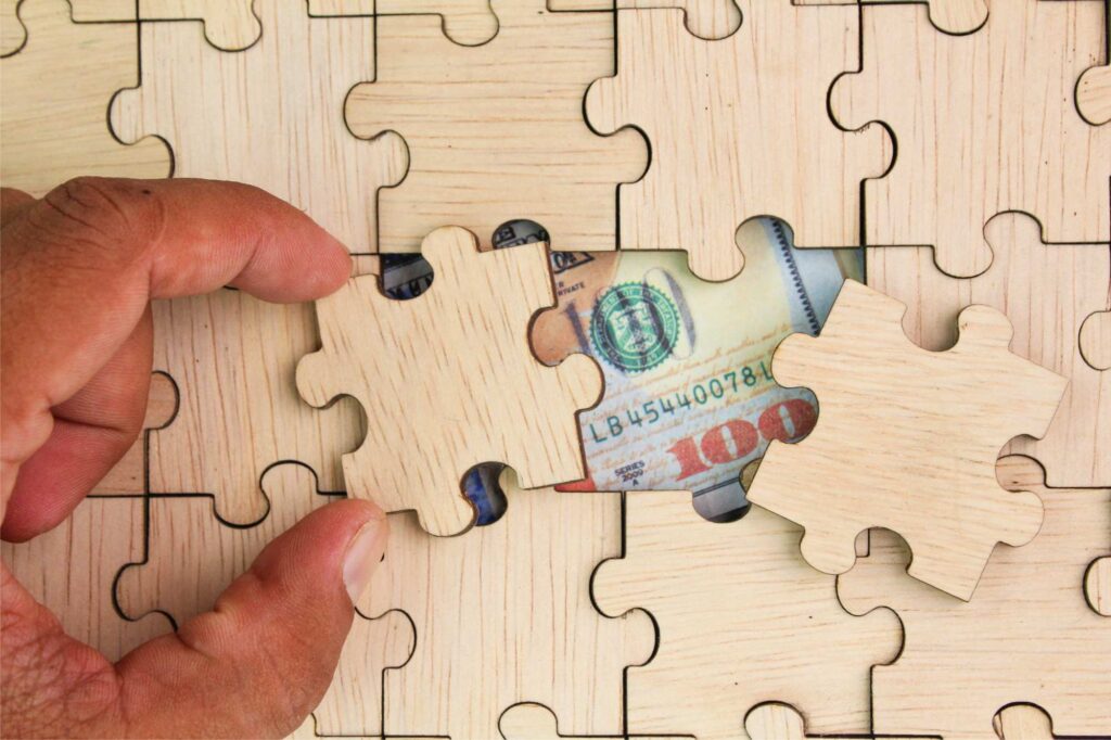 Hand placing a puzzle piece with money into a puzzle, illustrating contract profitability and connected business operations